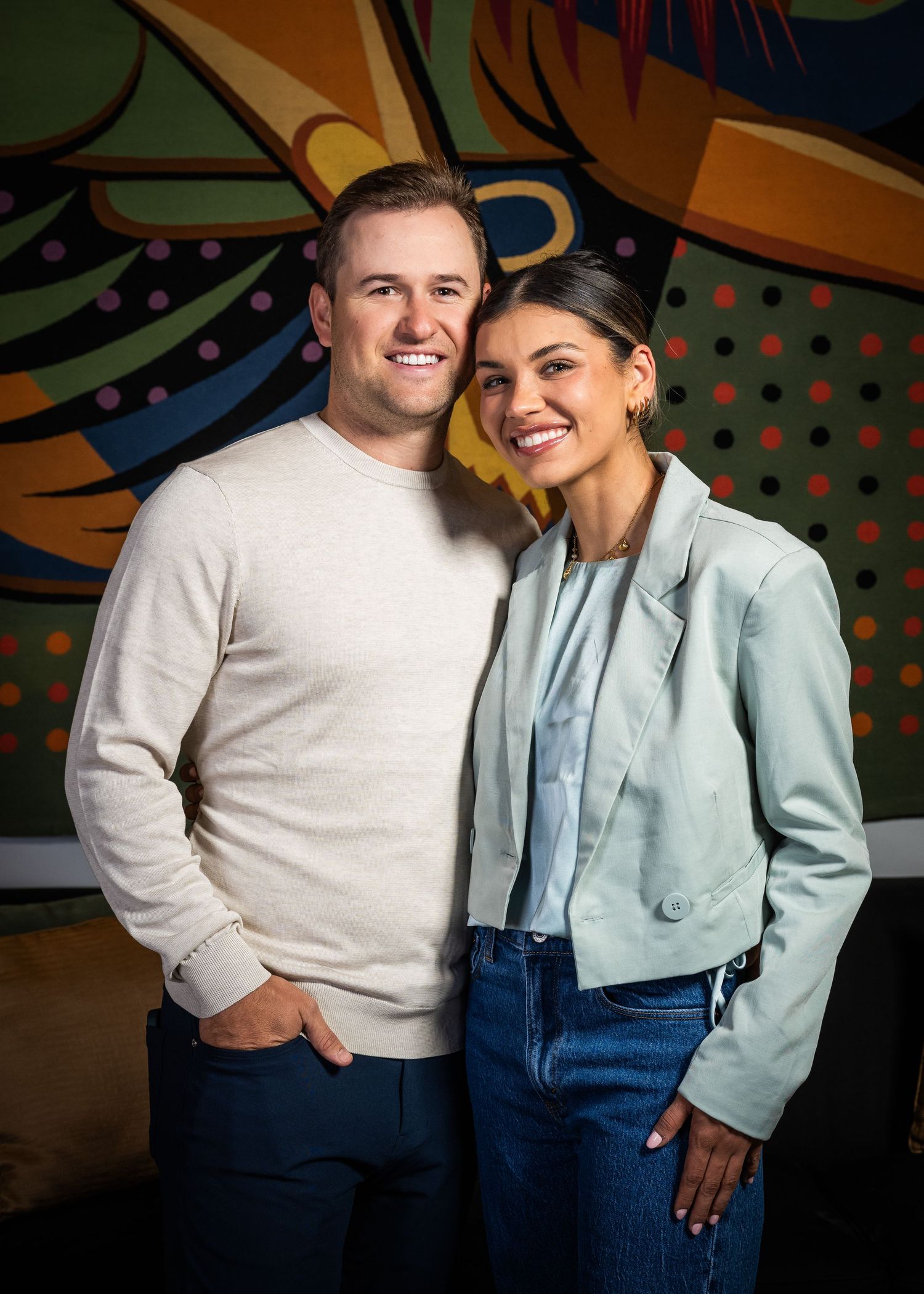 A smiling couple poses together in front of a colorful mural, dressed in casual smart attire.