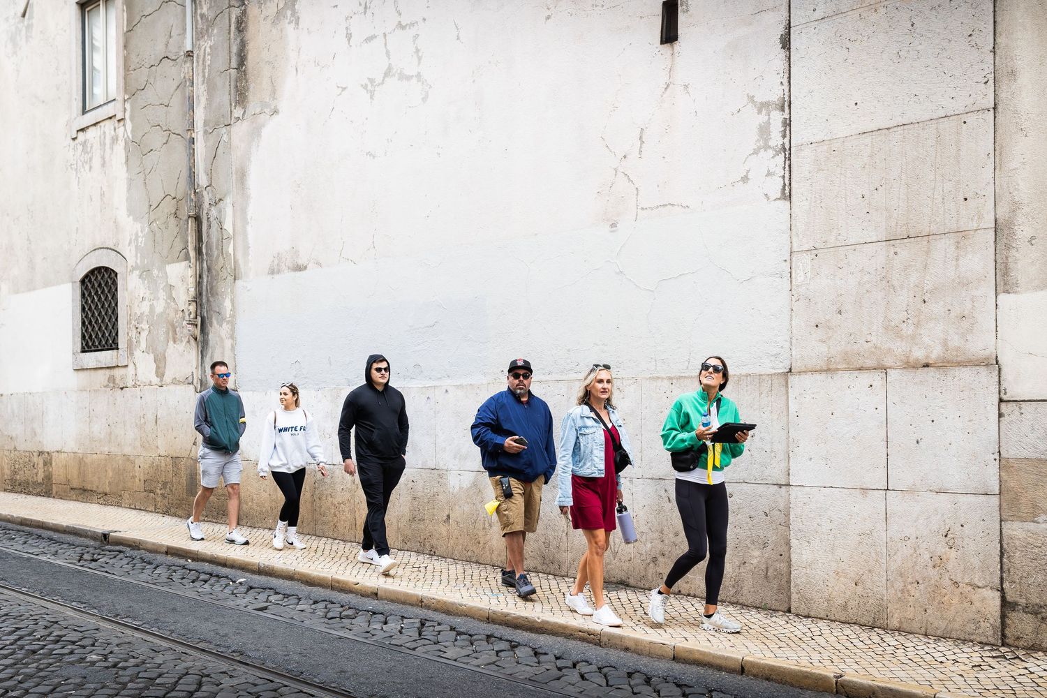 A group of tourists stands along an old stone building wall on a narrow Lisbon street.