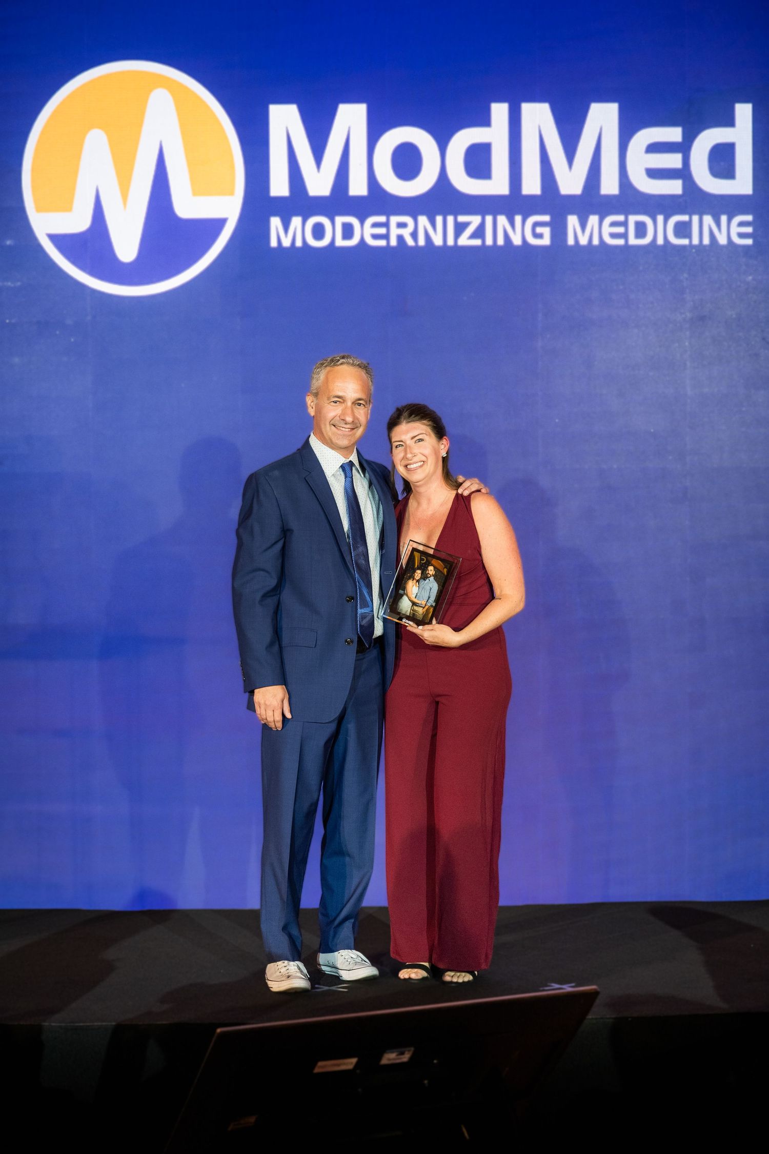 Two people posing with award in front of ModMed Modernizing Medicine branded backdrop.