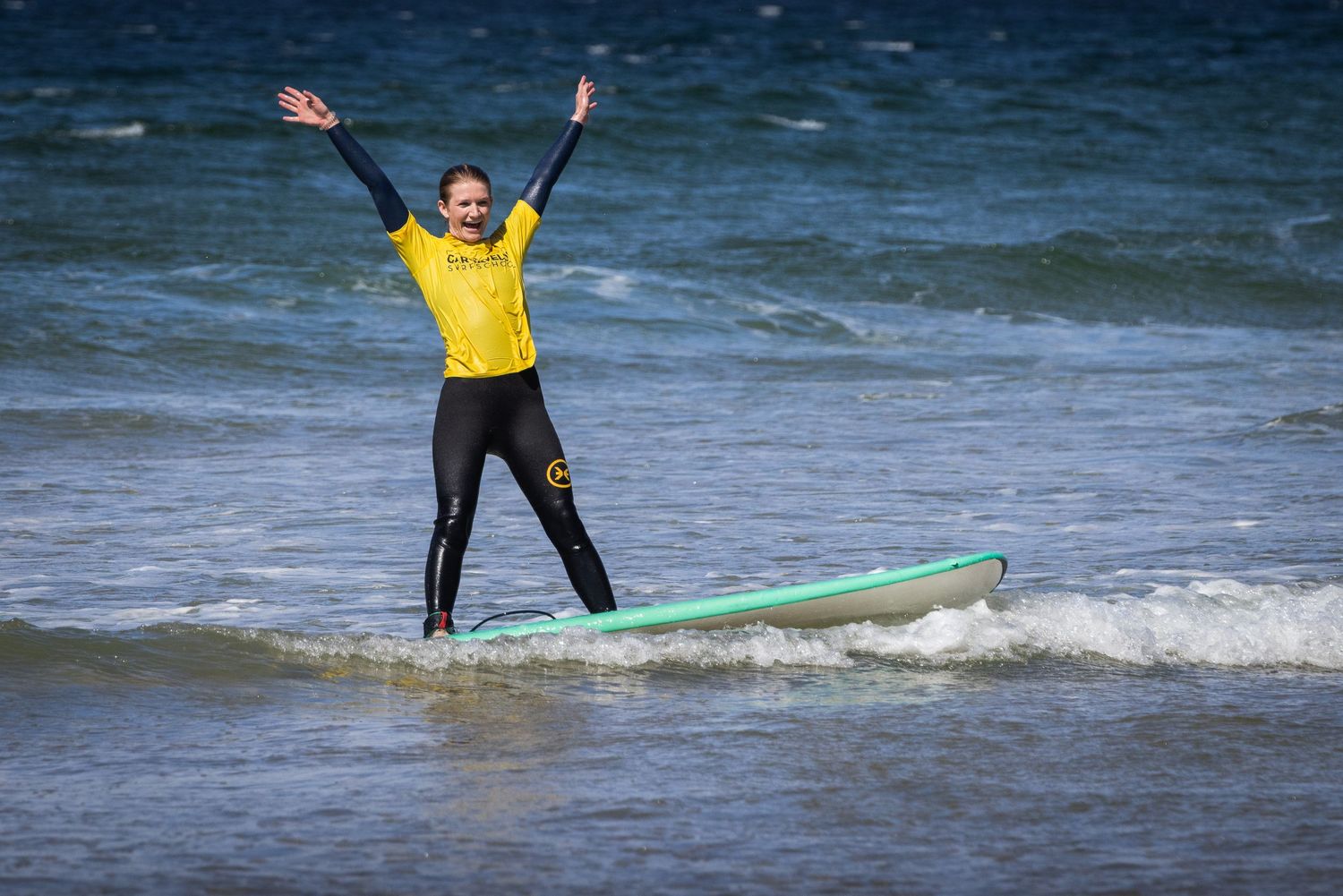 Excited beginner surfer celebrating with arms raised while riding a wave on a teal surfboard.