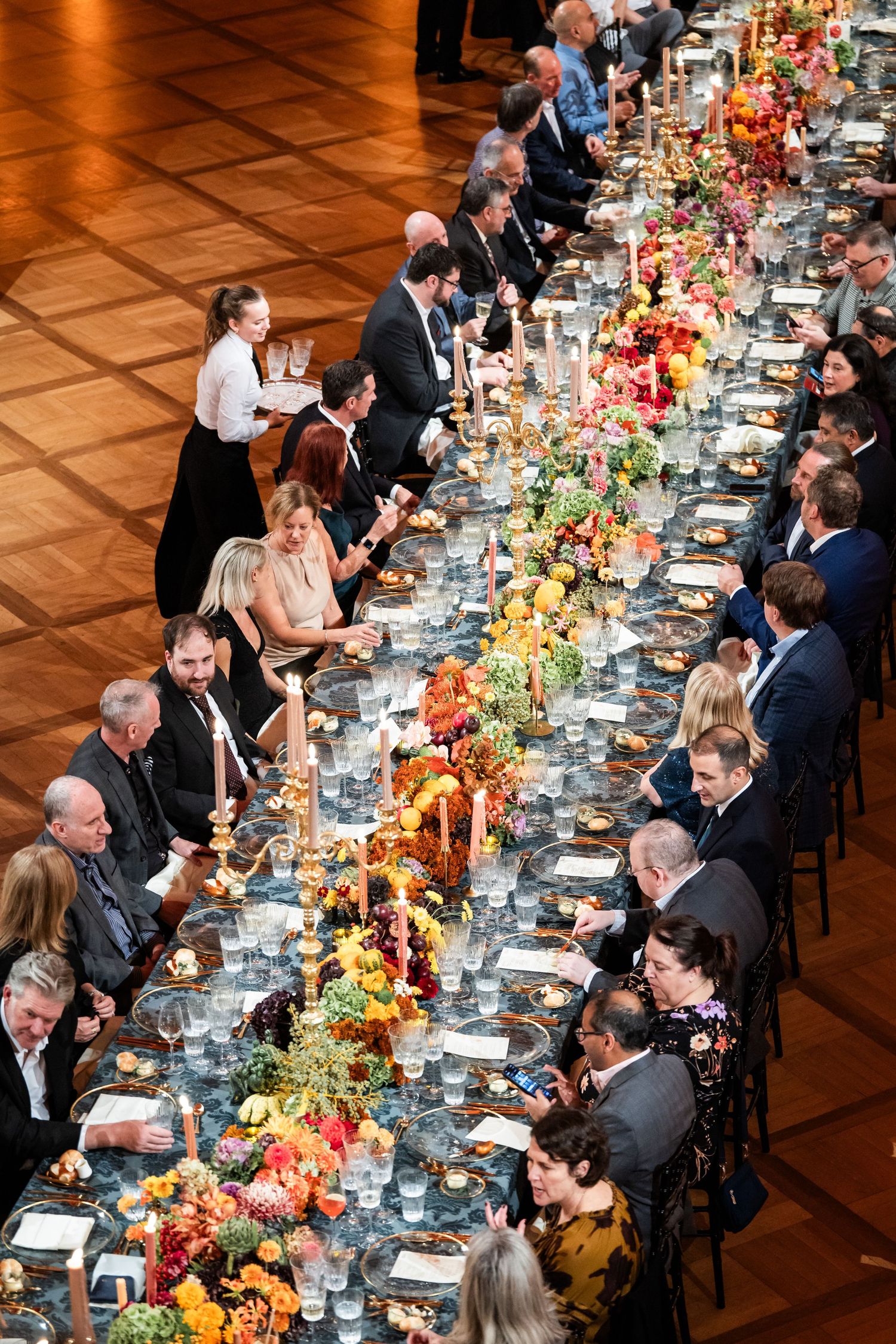 Aerial view of guests dining at a long banquet table adorned with colorful floral arrangements and candles at a formal gala.