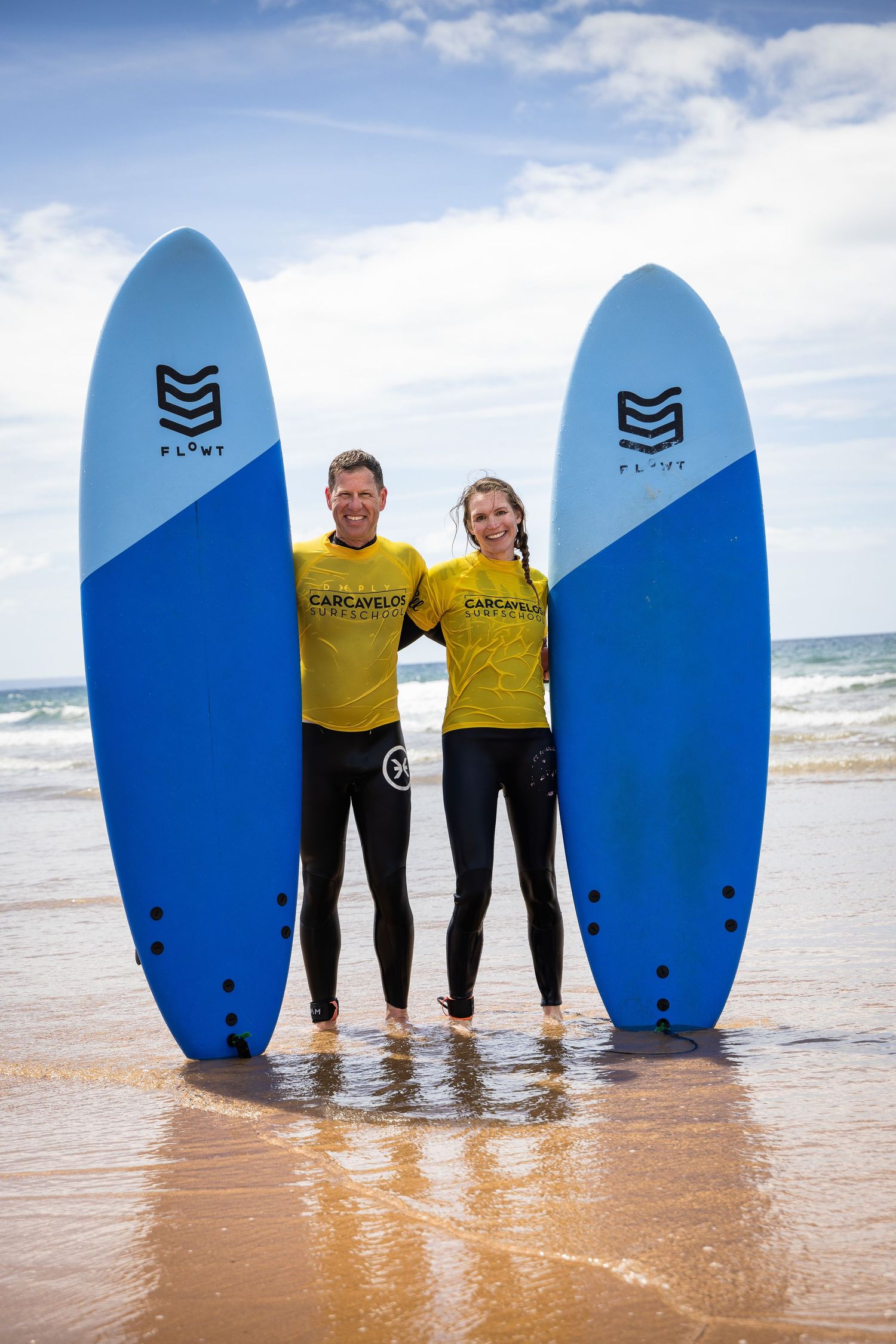 Two surfers in yellow Carcavelos wetsuits posing with blue Flyt surfboards on a sunny beach.