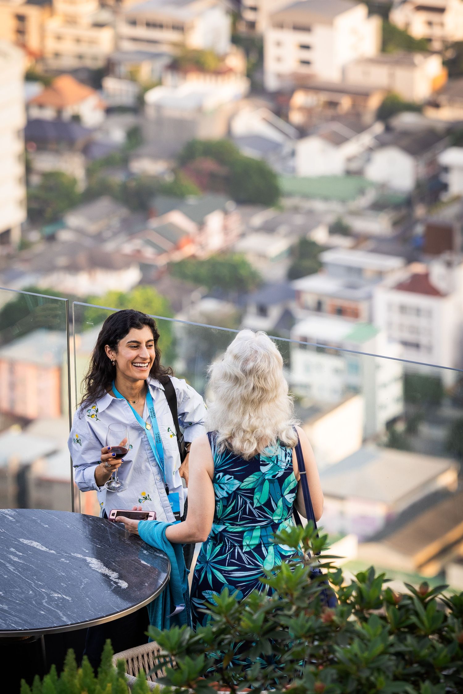 Two women conversing at a rooftop bar with a panoramic aerial view of a city below.