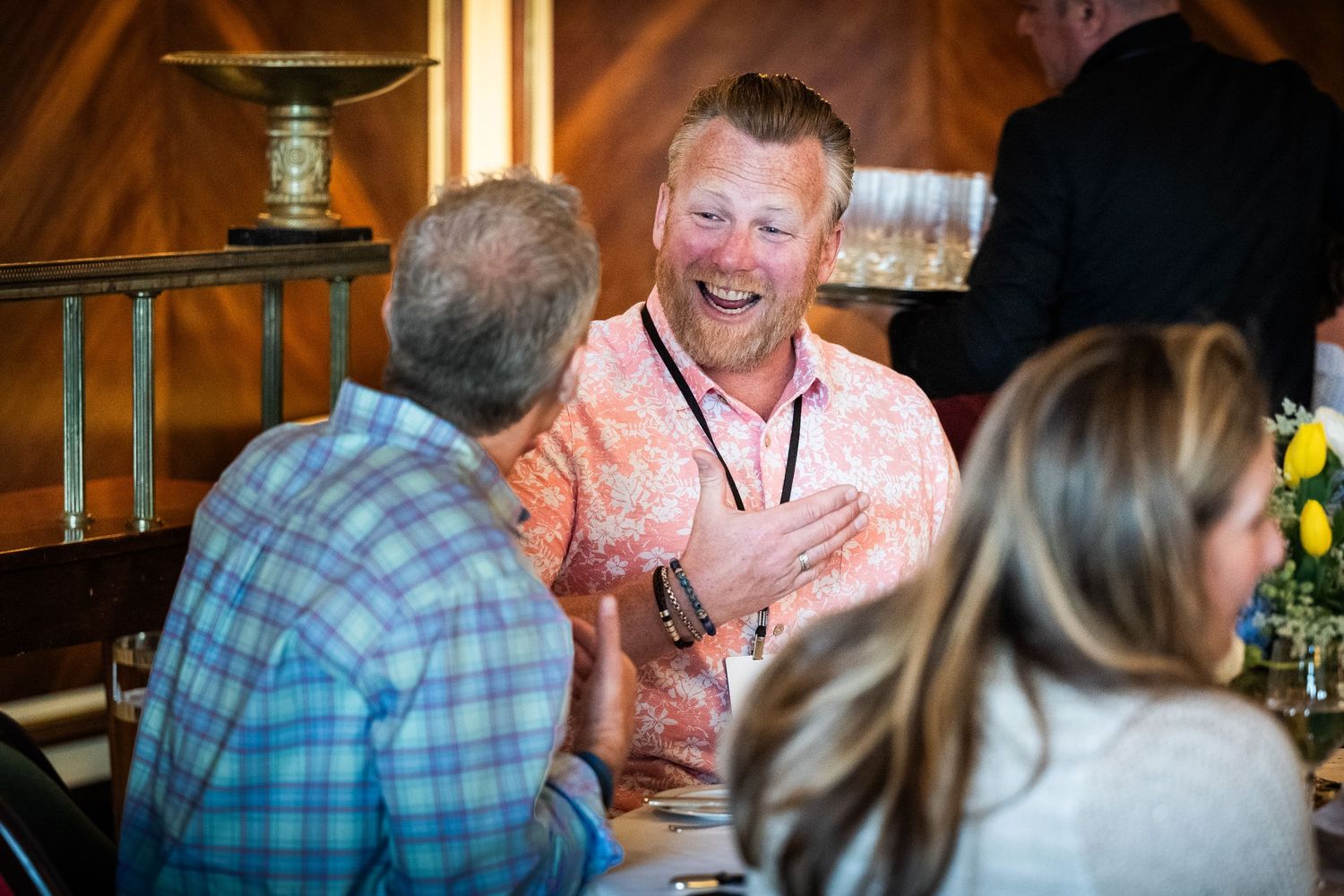 A man in a pink floral shirt laughs heartily while socializing at a casual gathering.