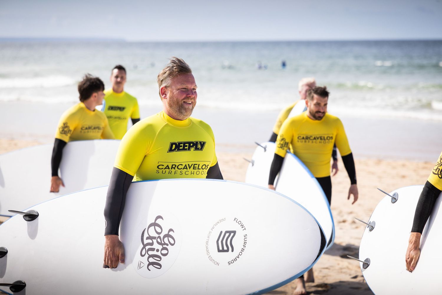 Group of smiling surfers in yellow Carcavelos wetsuits carrying white surfboards on a sunny beach.