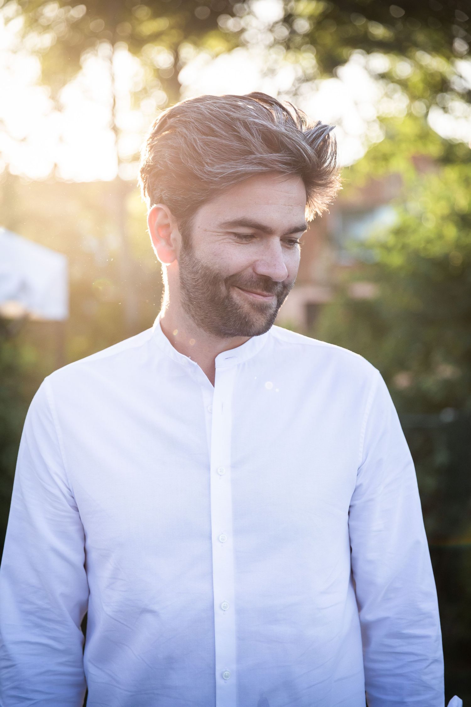 Smiling bearded man in a white shirt enjoying a sunny outdoor setting with green trees behind him.