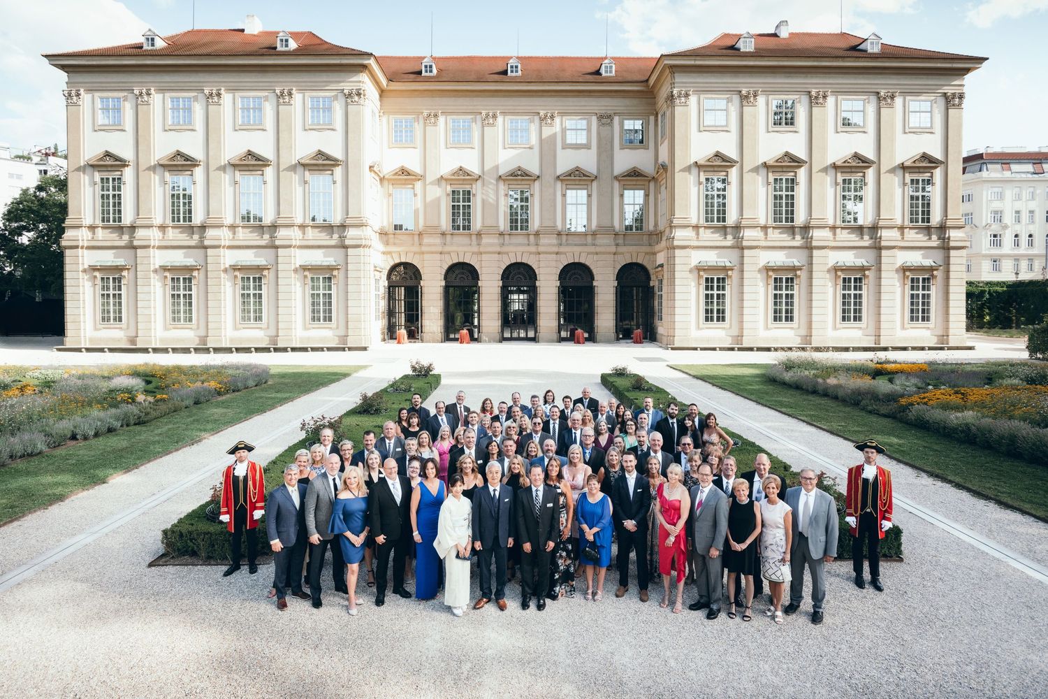 Large group of professionals posing in front of an elegant European palace building surrounded by manicured gardens.