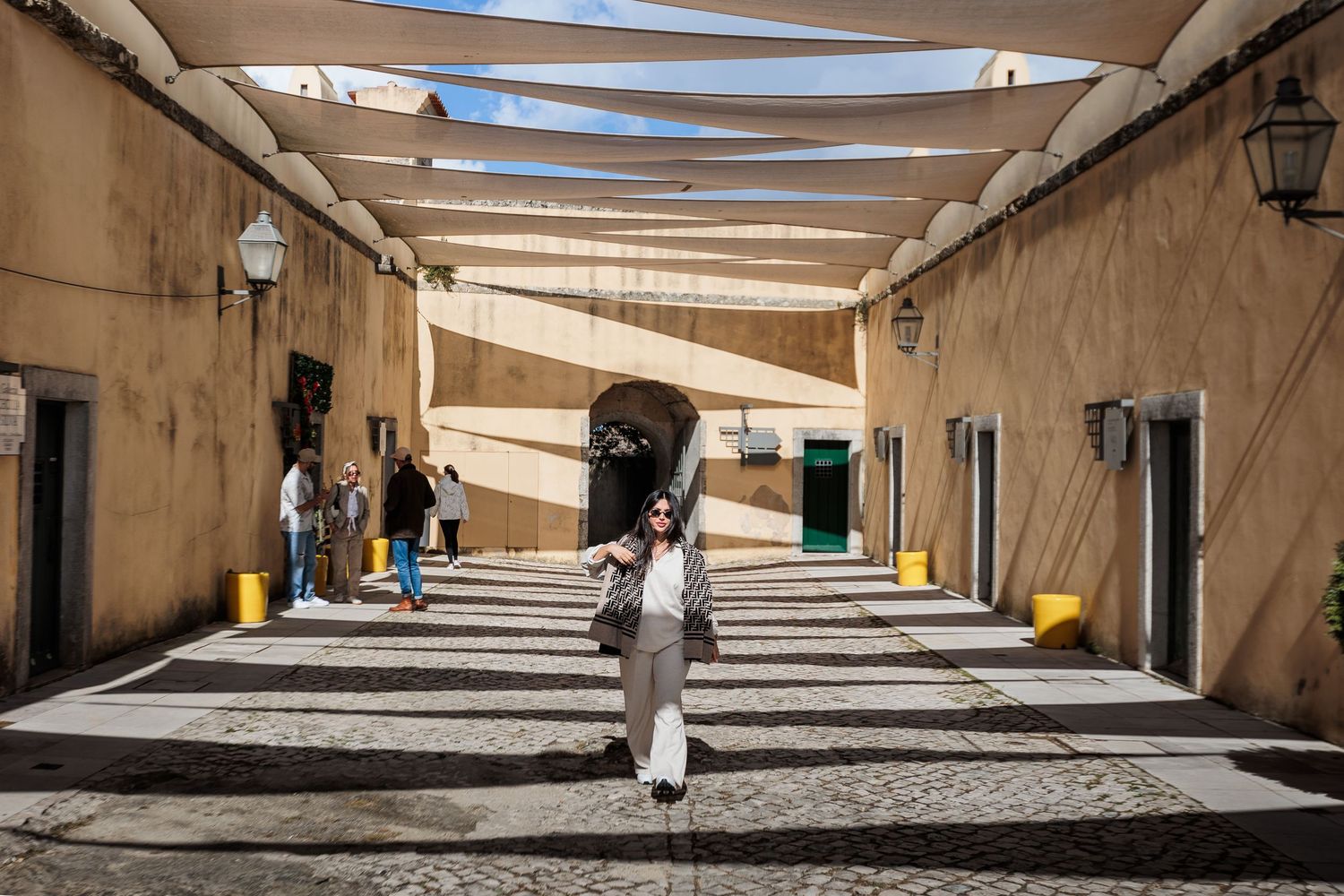 Woman walking through a sunlit covered alleyway with white shade sails in a historic Mediterranean town.