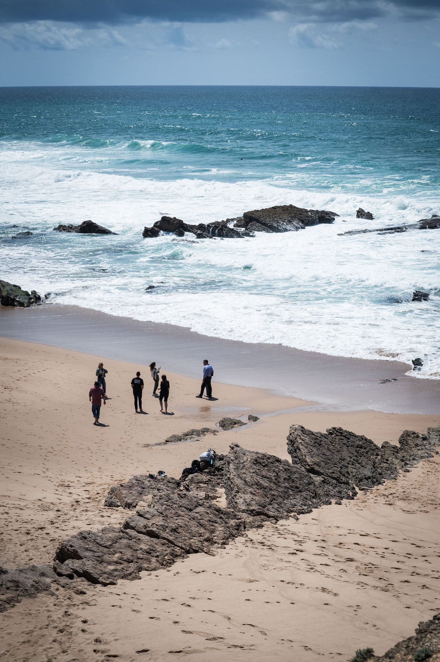 Group of people standing on a sandy Atlantic Ocean beach with rocky shoreline and waves.