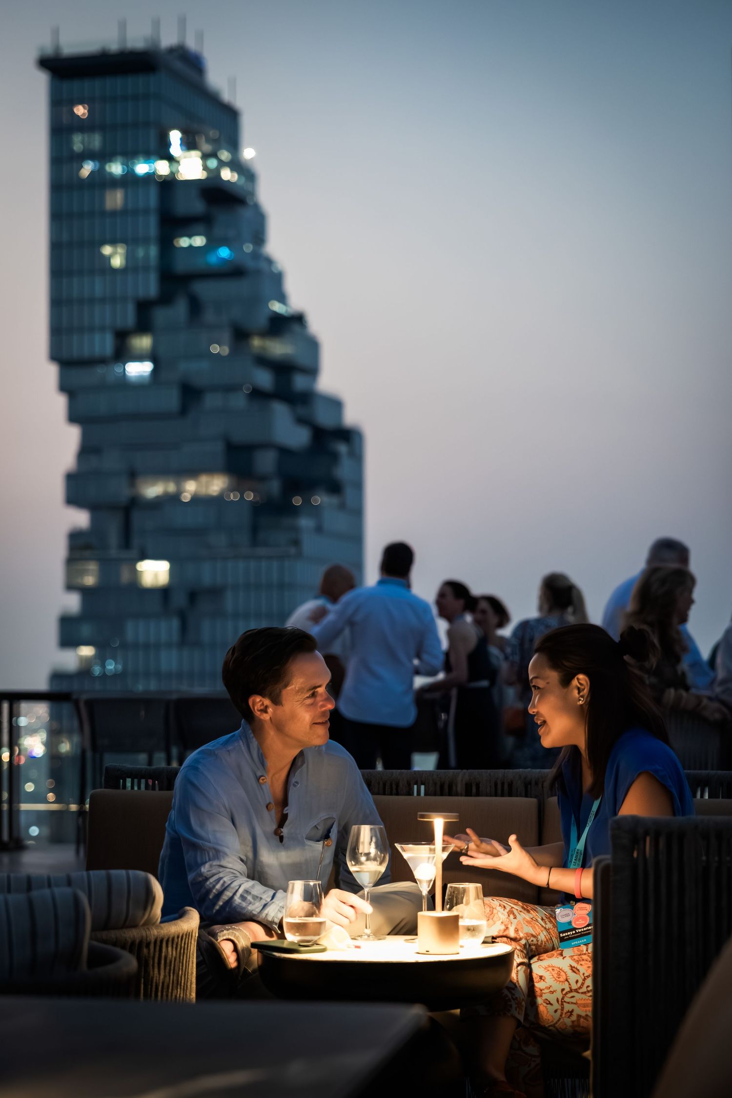 Couple enjoying a romantic candlelit dinner on a rooftop bar with a stunning Bangkok skyline at dusk.