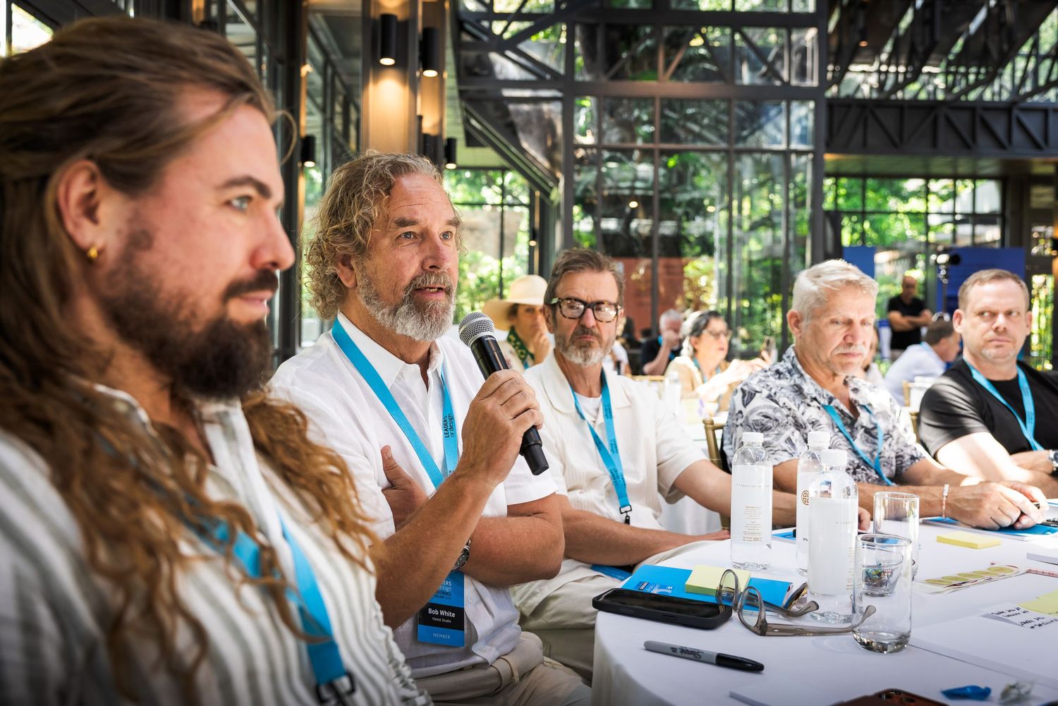 Panel of men speaking into a microphone at an outdoor conference table during a daytime event.