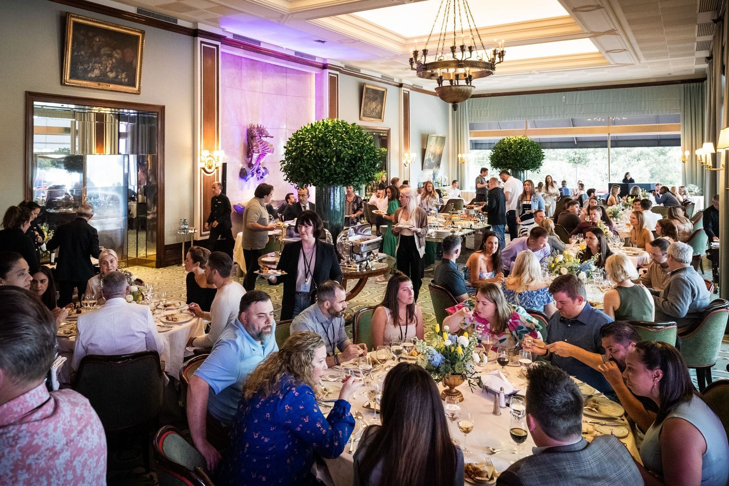 A large crowd of elegantly dressed guests dine together in a beautifully decorated banquet hall.