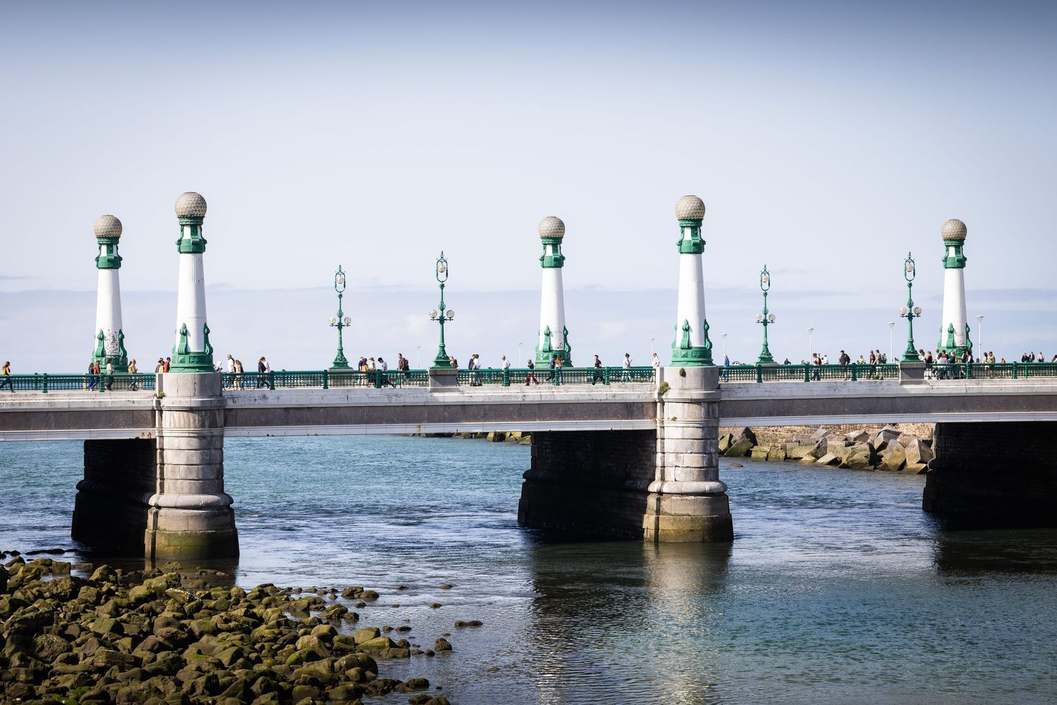 Historic pier with ornate green and white lamp posts extending over calm coastal waters under a clear sky.