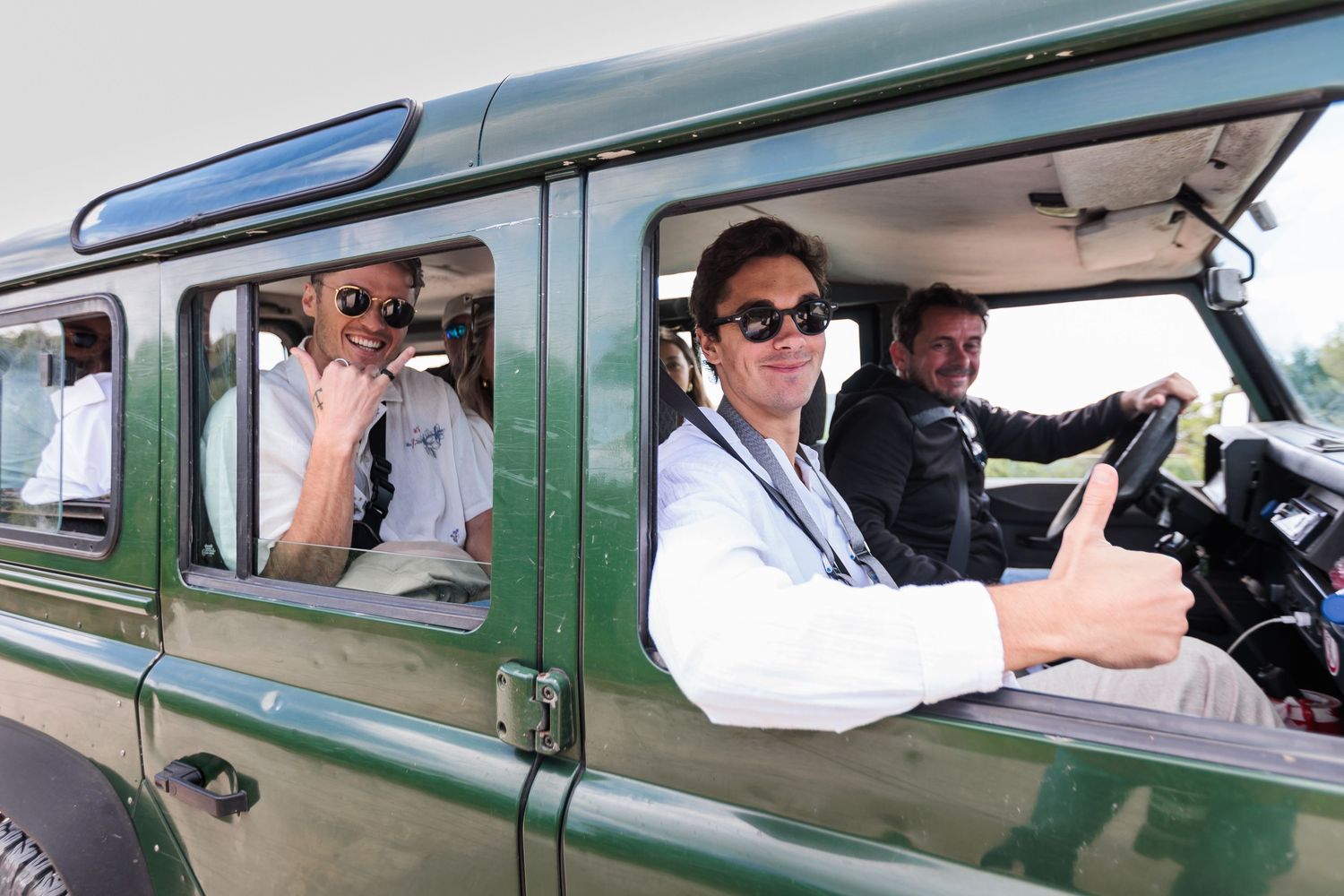 Three men smiling and giving thumbs up inside a classic green Land Rover Defender on a sunny day.