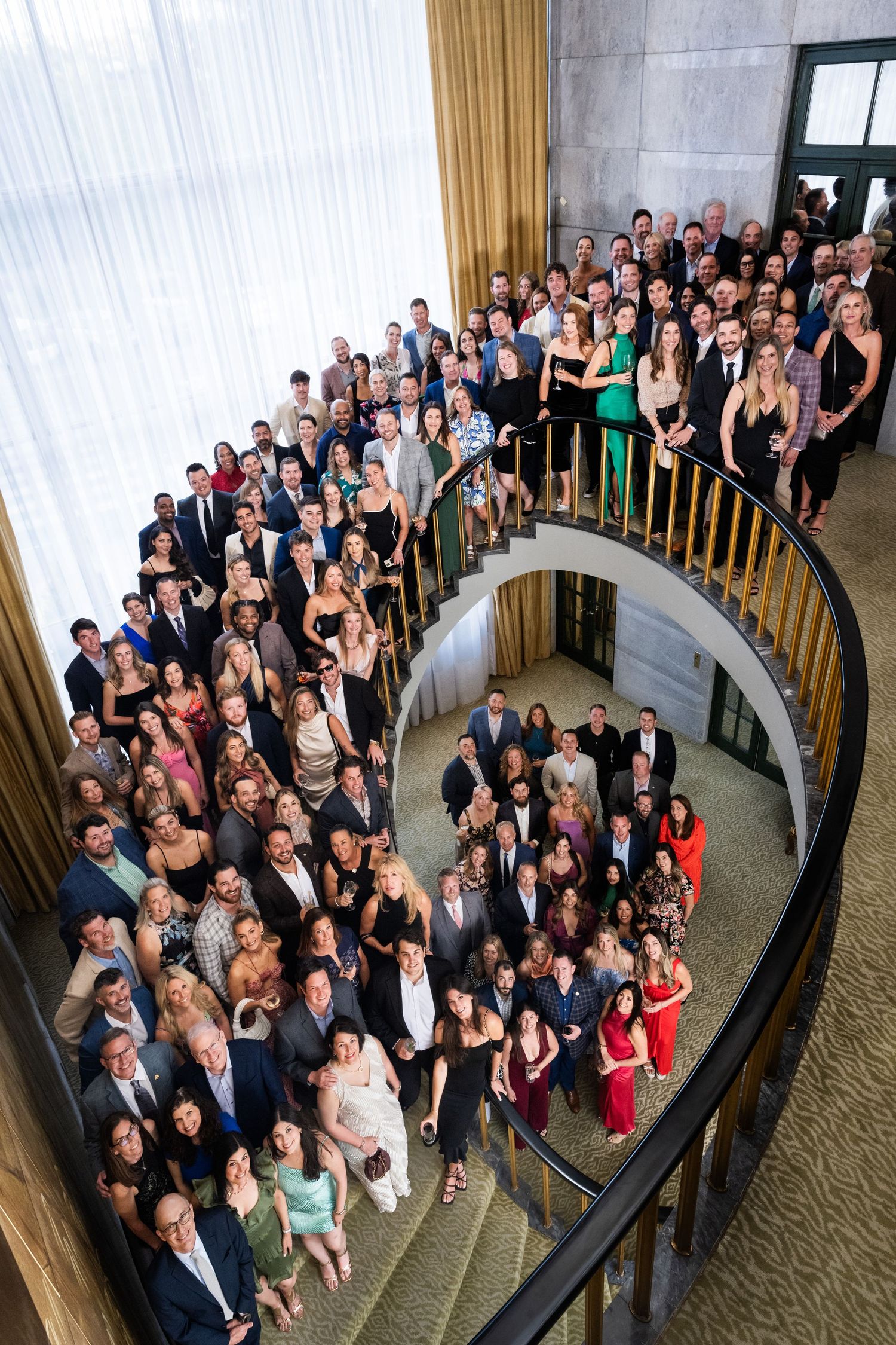 Large elegantly dressed group posing on a grand spiral staircase at a formal corporate event.