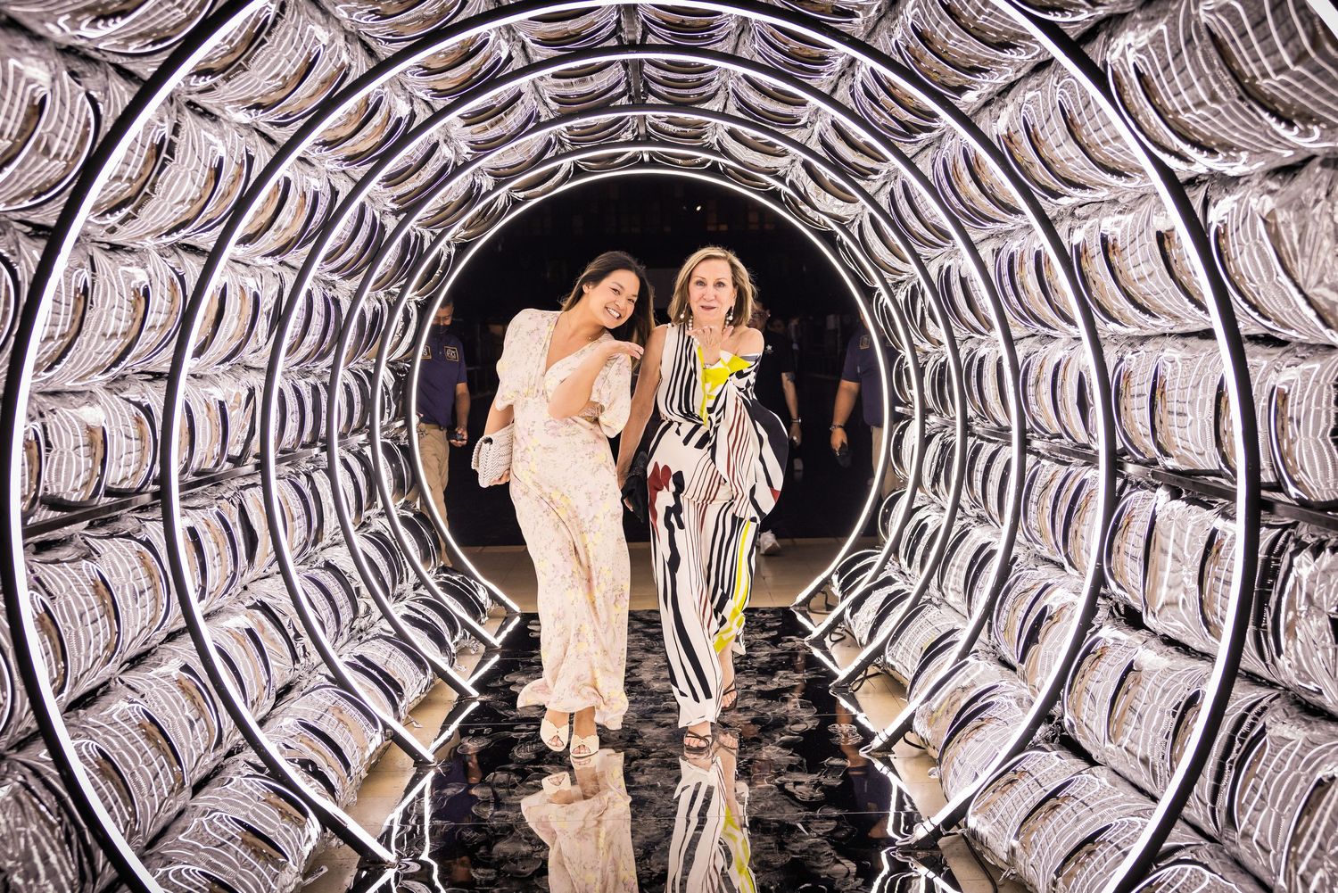 Two women in glamorous outfits walking through a dazzling mirrored tunnel installation.