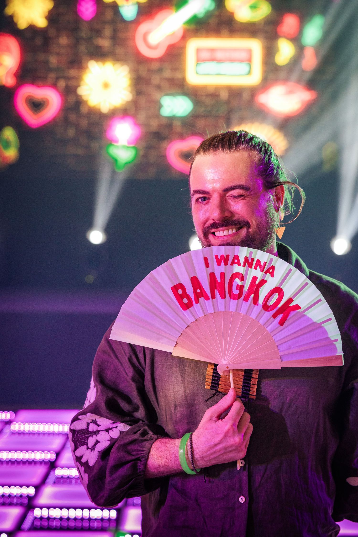 Smiling man holding a decorative fan reading I Wanna Bangkok at a neon-lit venue.
