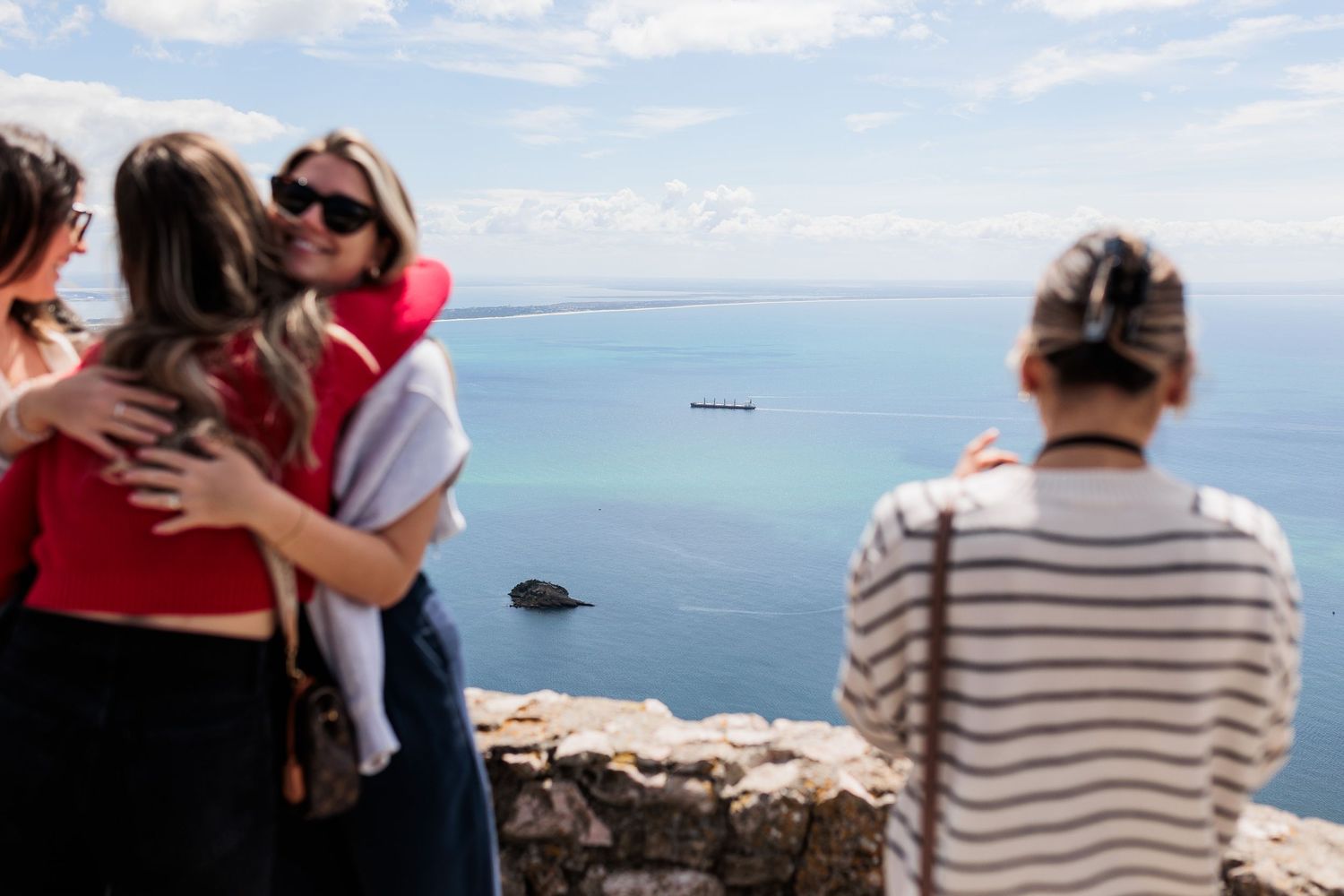 Two women embracing at a scenic coastal viewpoint while a friend photographs them with ocean backdrop.