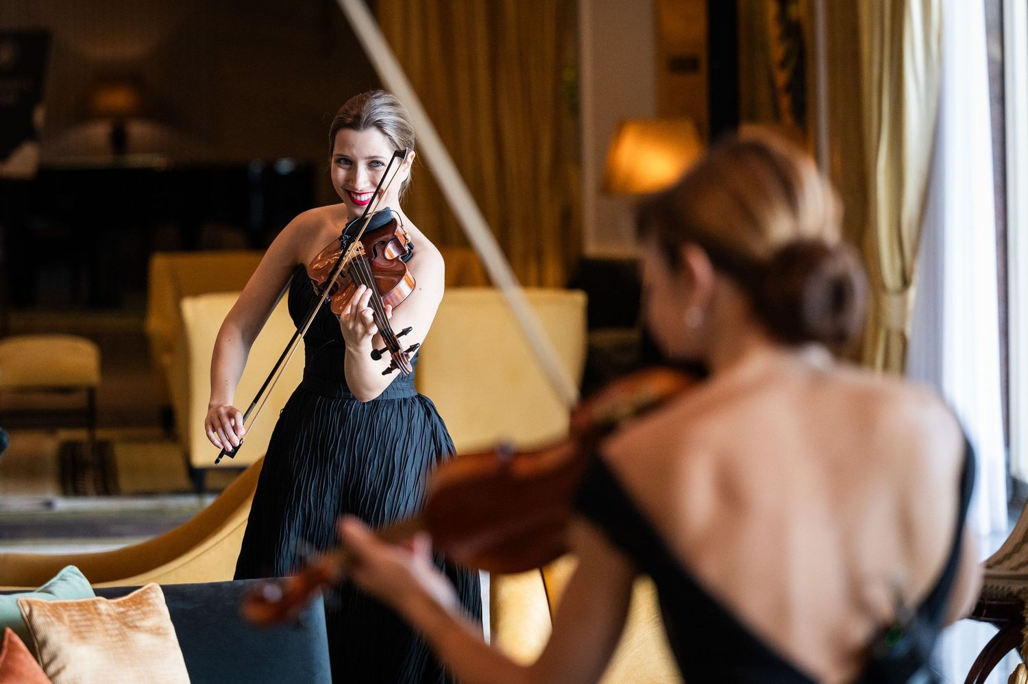 Two female violinists in black dresses performing in an elegant upscale hotel lounge setting.