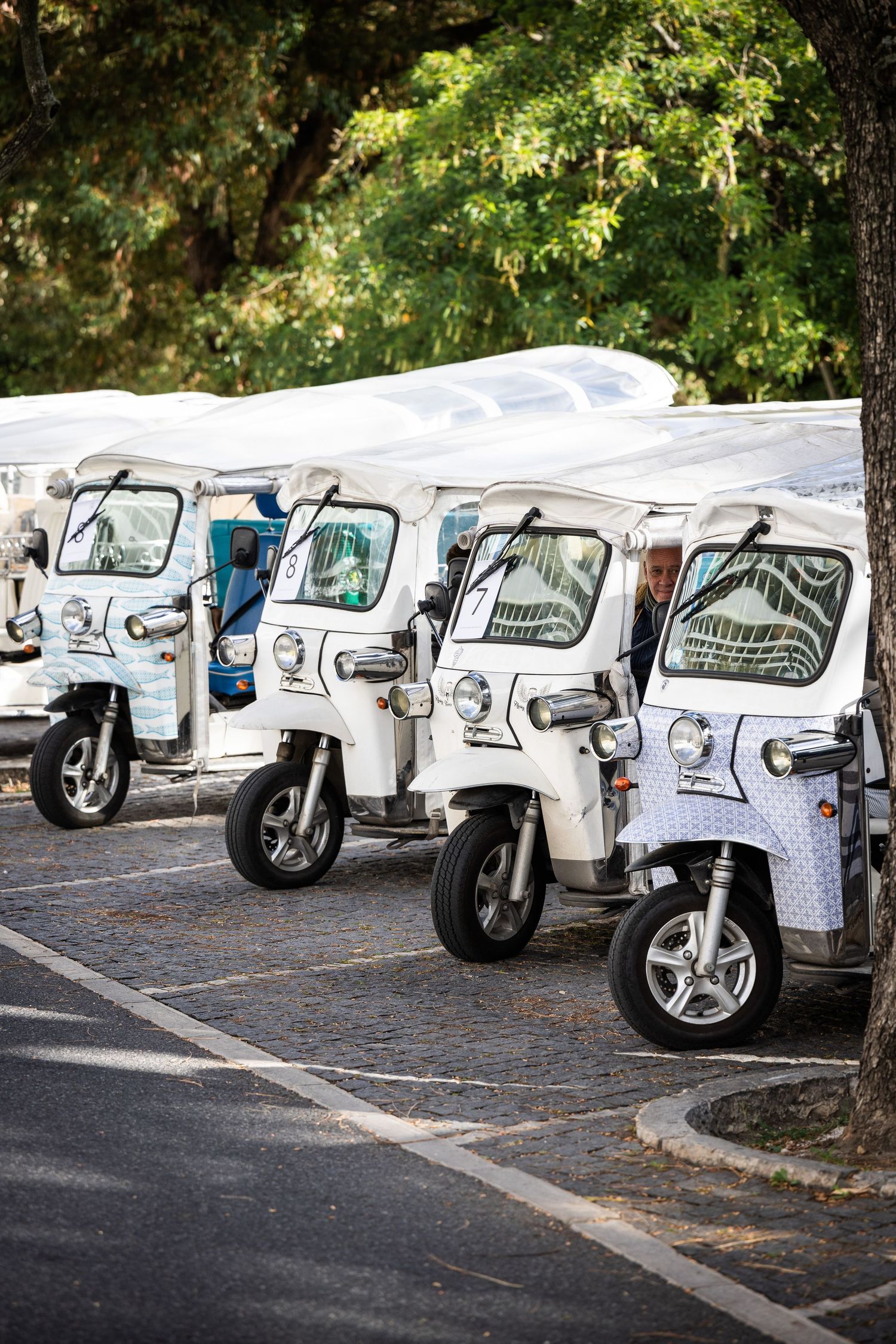 A row of white tuk-tuk electric vehicles parked on cobblestone street under green trees.