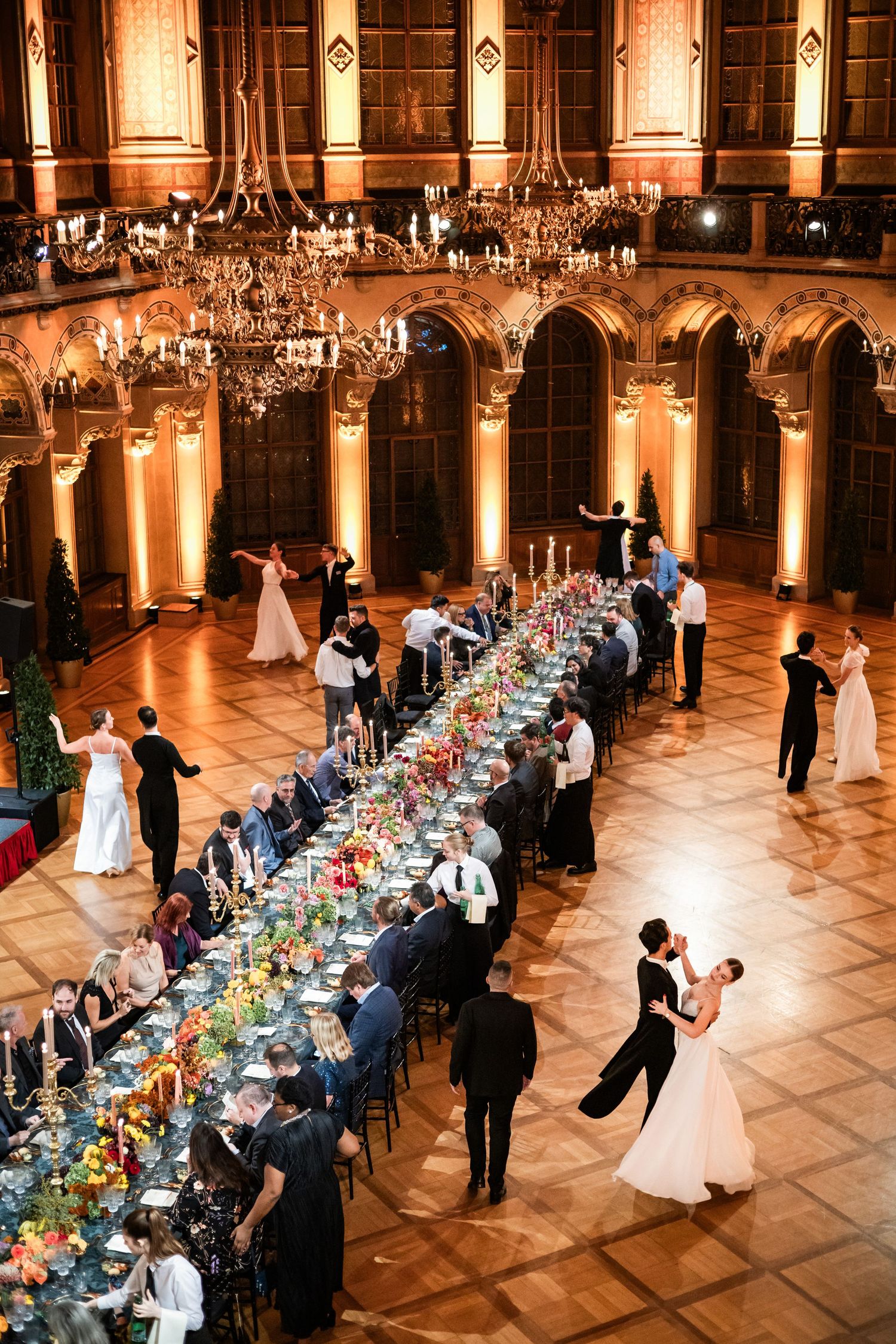 Elegant wedding reception with couples dancing beside a long floral table in a grand chandelier-lit ballroom.