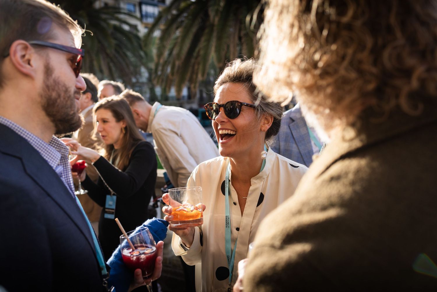 Woman laughing joyfully while holding food and drinks at a sunny outdoor social event.