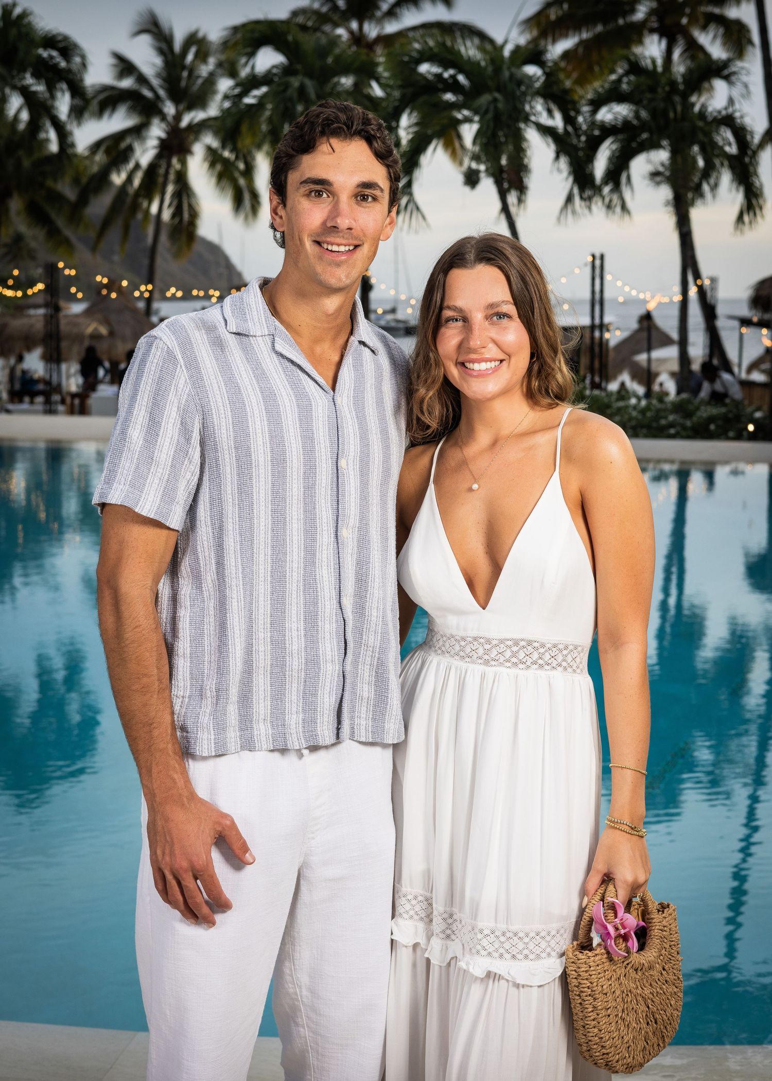 Couple dressed in white posing together poolside at a tropical resort during sunset.