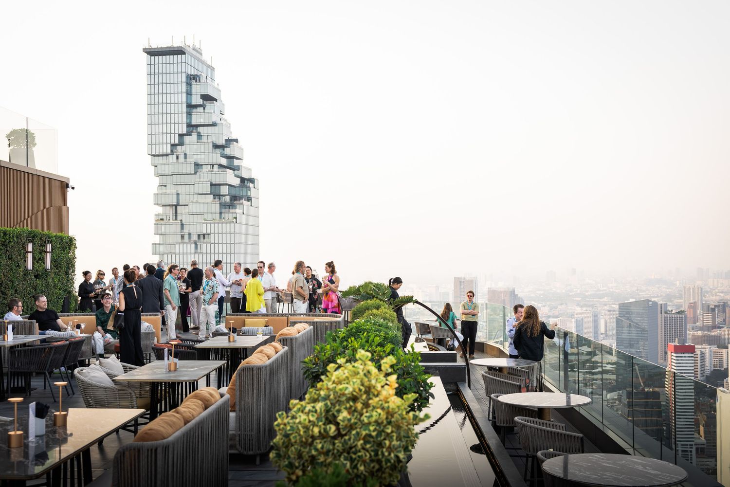 Guests socializing on a luxurious rooftop bar overlooking the Bangkok cityscape at sunset.