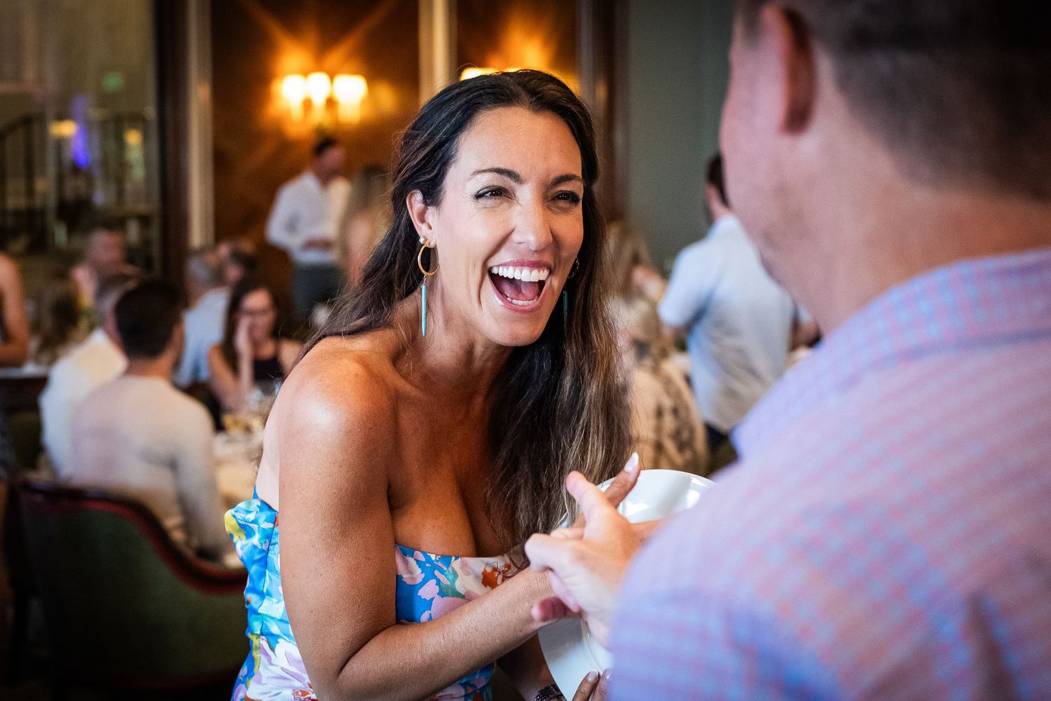 A woman laughs joyfully at an elegant restaurant event while engaging in conversation with another guest.