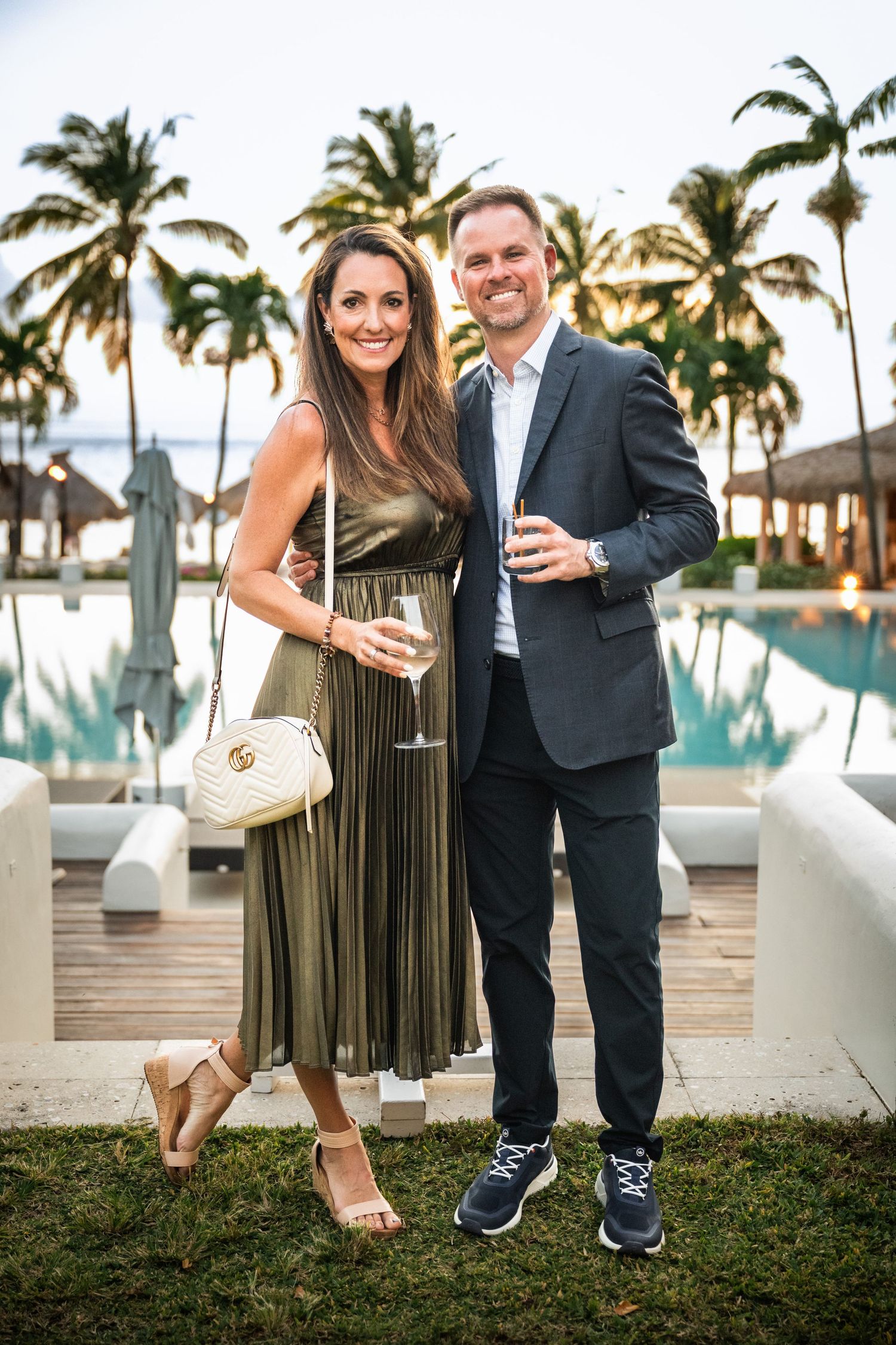 Elegant couple holding champagne glasses posing by a resort pool surrounded by palm trees.