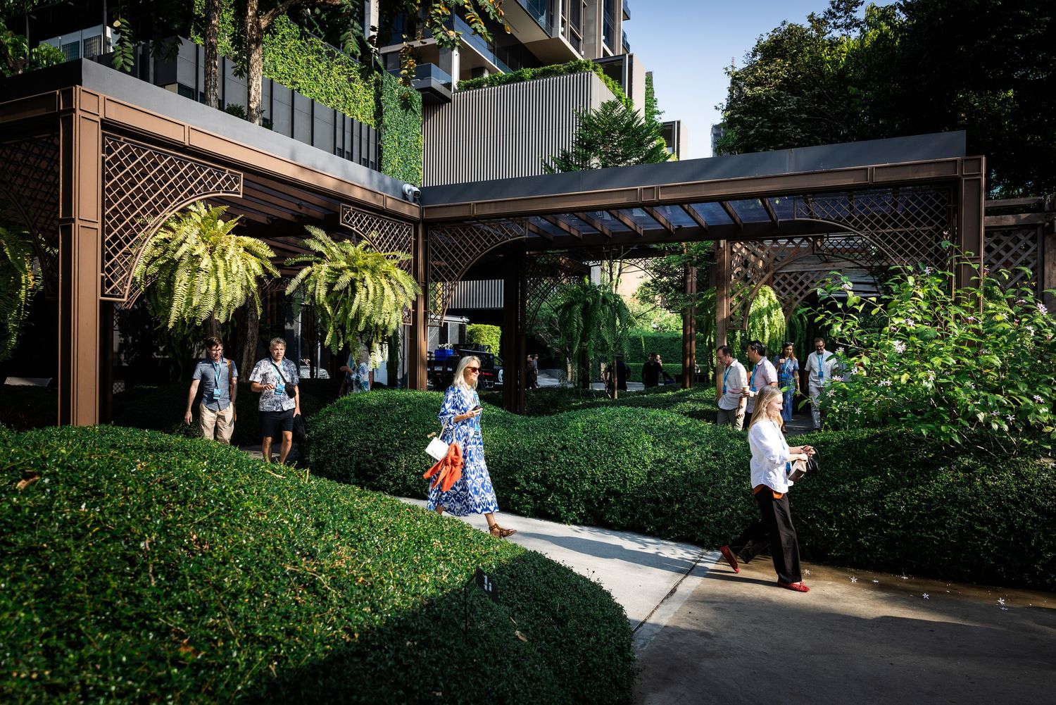 People walking through a lush green garden courtyard with wooden pergolas and tropical plants.