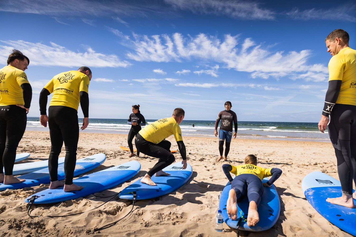 Surf instructor teaching beginners proper surfboard techniques on a sandy beach with blue boards.