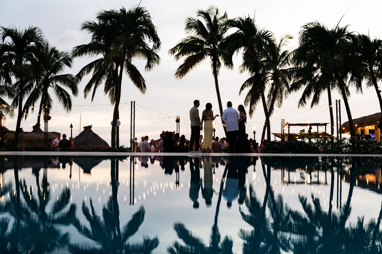 Silhouettes of people standing by a resort pool at sunset with tall palm trees reflecting in water.