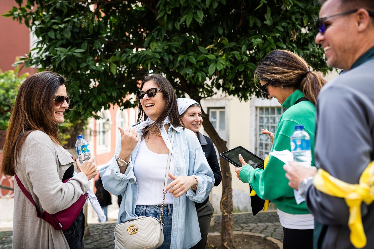 Group of tourists smiling and chatting outdoors near green trees on a sunny day.