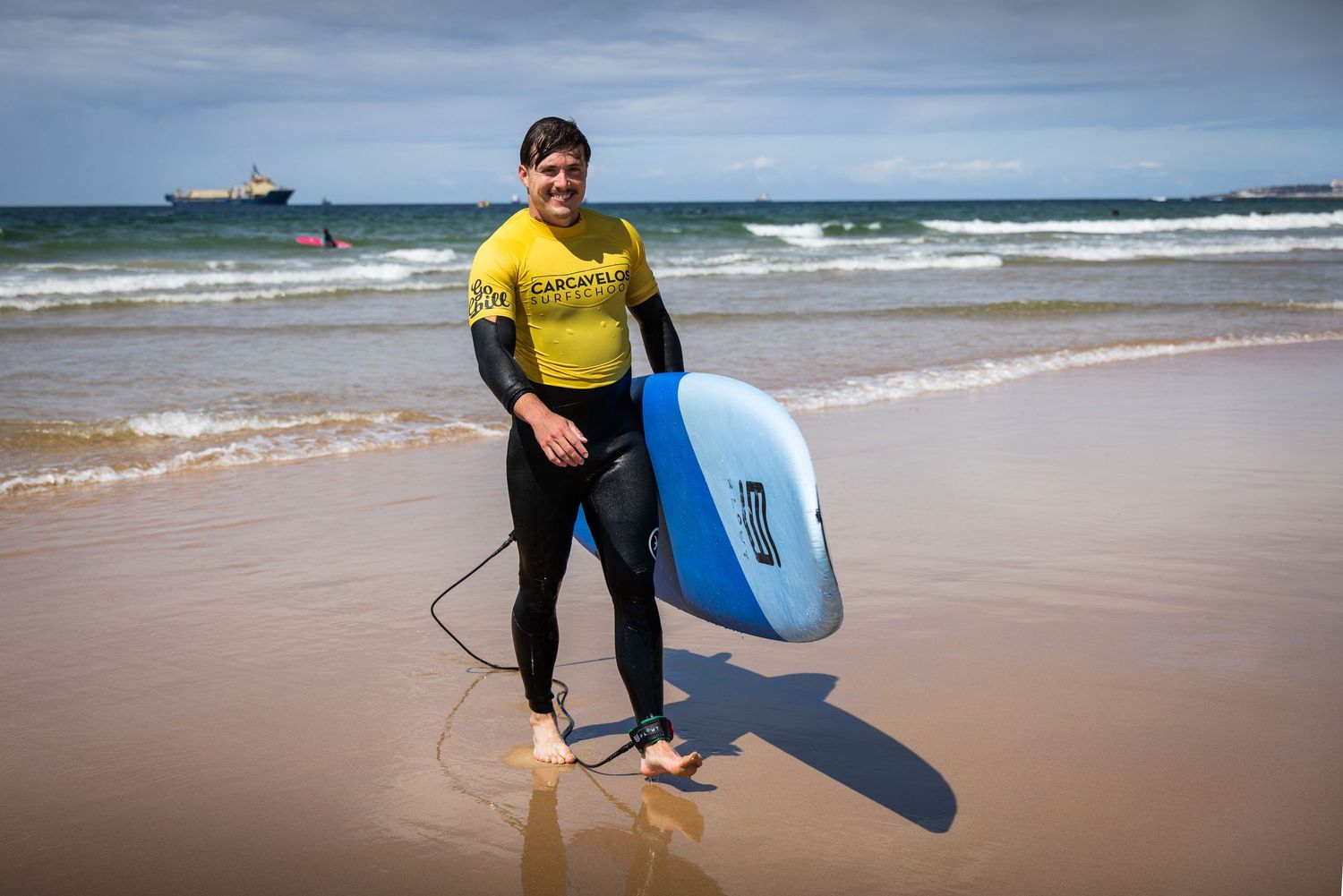 Male surfer in yellow Carcavelos wetsuit holding a blue bodyboard on a sandy beach shoreline.
