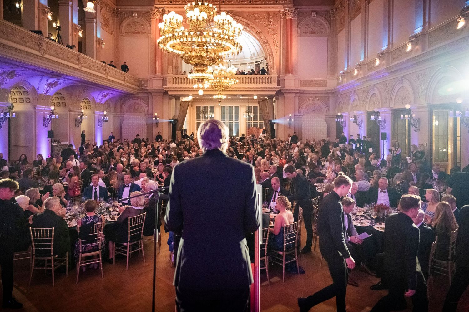 Speaker addresses a large elegant banquet hall filled with guests seated at round dining tables.