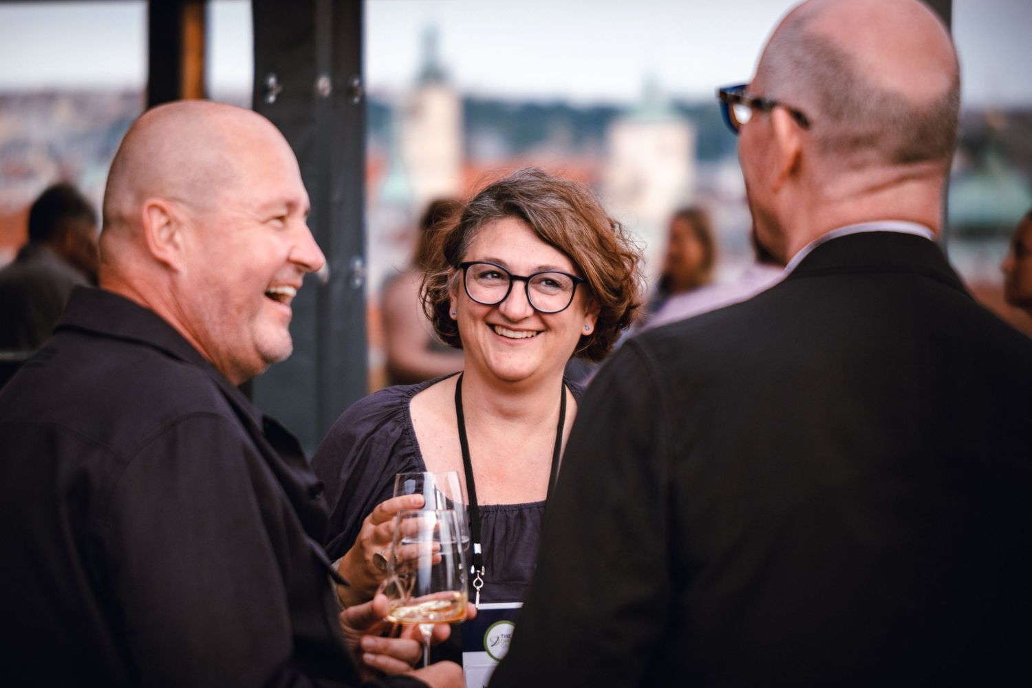 Three professionals networking and laughing at a rooftop corporate event while holding champagne glasses.