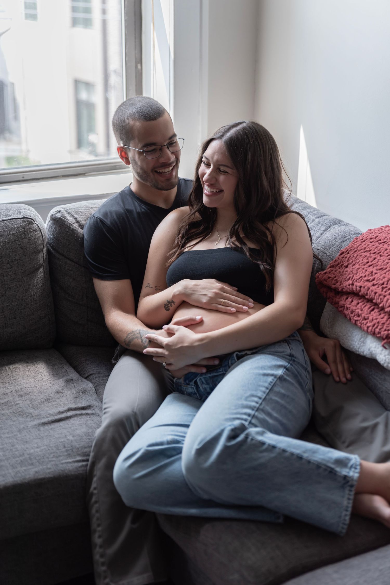 A couple sits on the couch together laughing and holding her belly during their maternity shoot in Edgewater, NJ