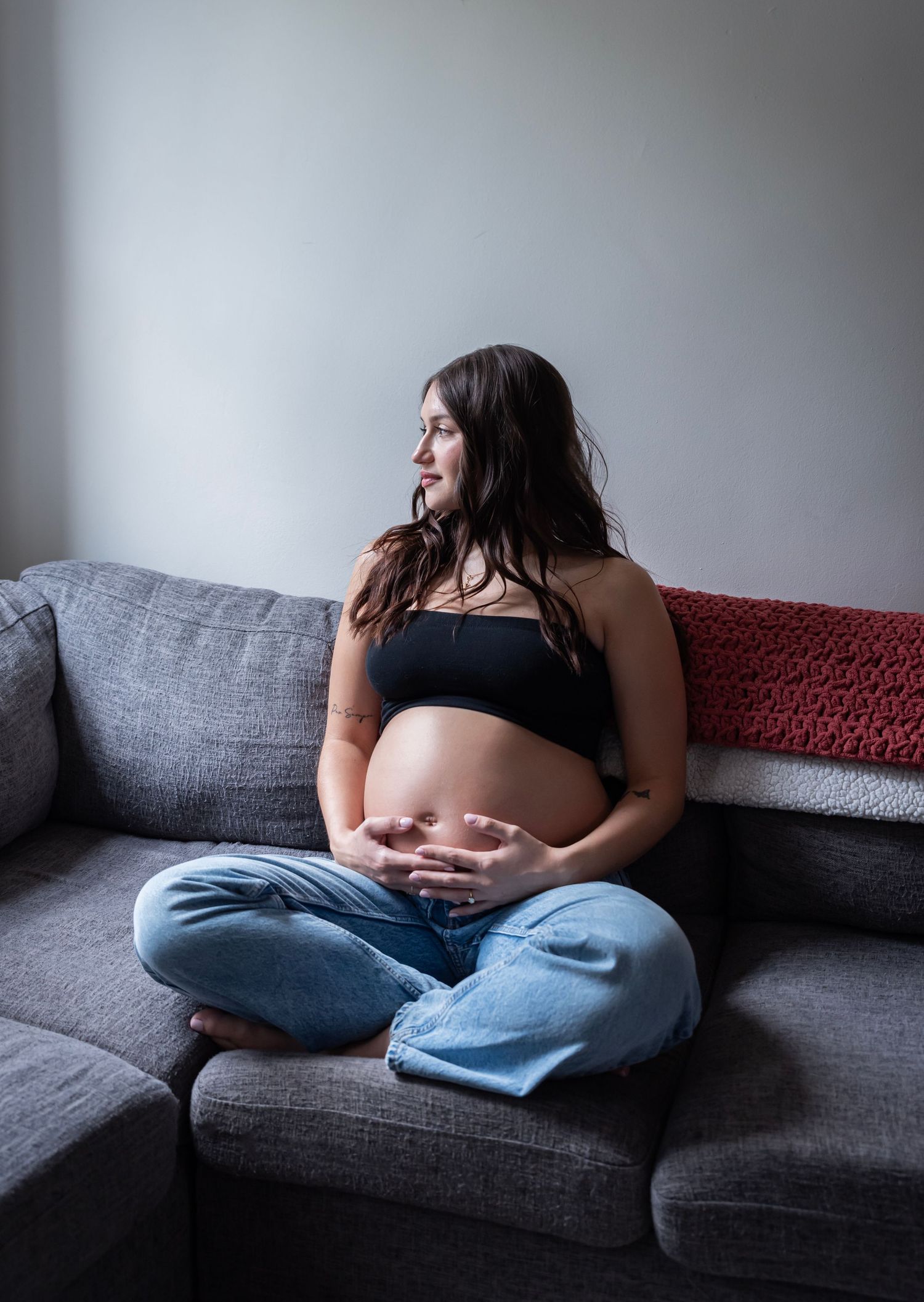 A pregnant woman sits on her couch holding her belly looking out the window during her maternity session in Edgewater, NJ