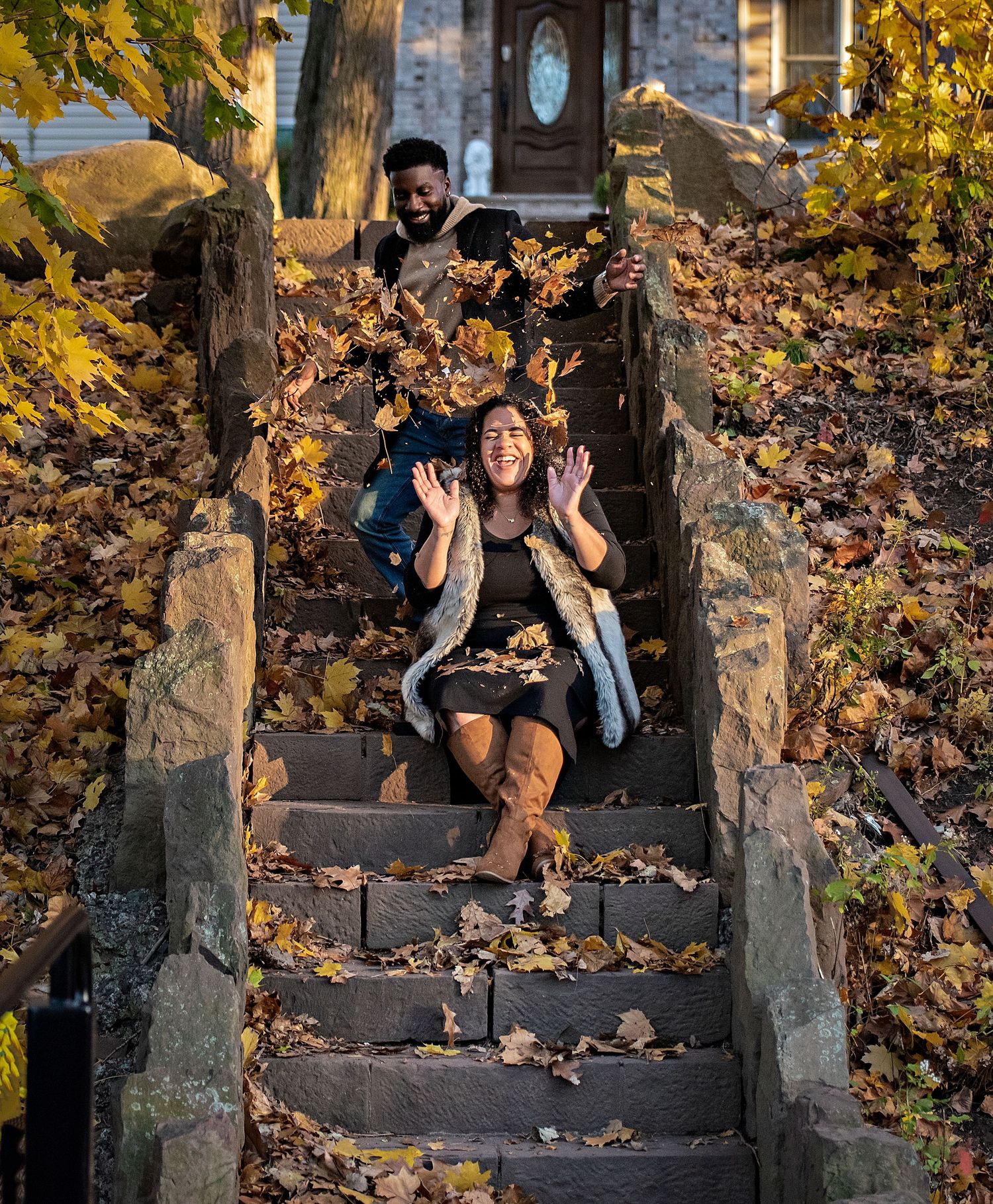 A couple during their maternity shoot has some fun and the father throws a pile of leaves onto the mom to be as she laughs in Nutley NJ