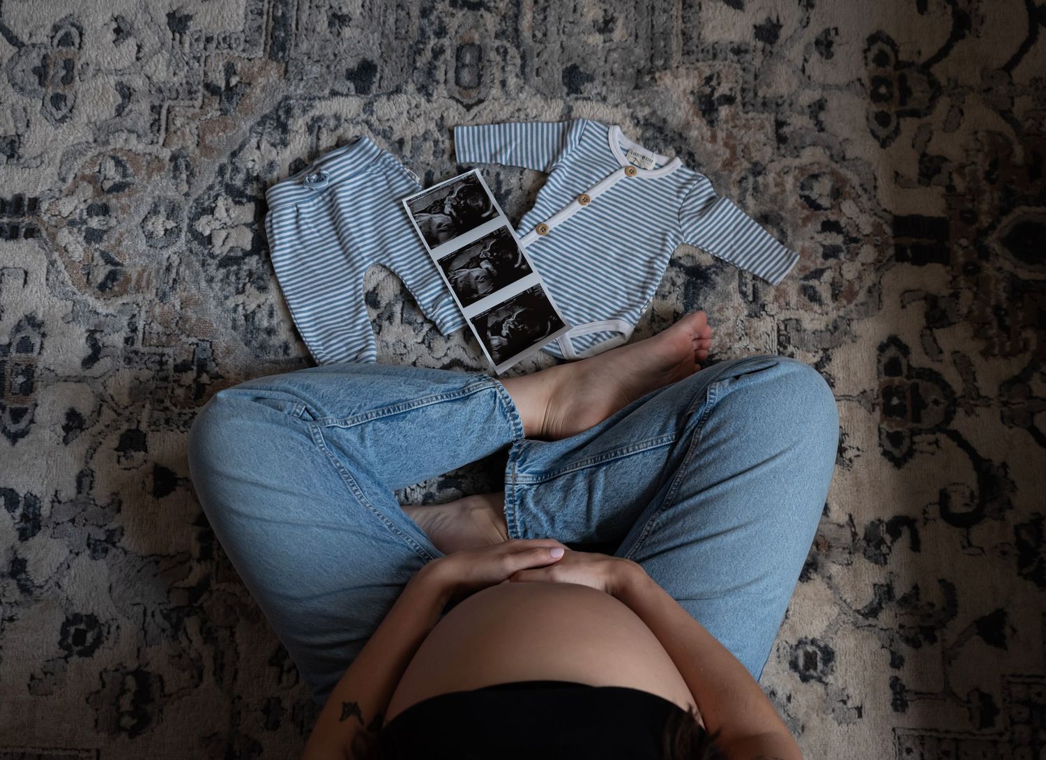A mother to be is sitting with a picture of a sonogram and babies clothing in her maternity photoshoot in Edgewater, NJ