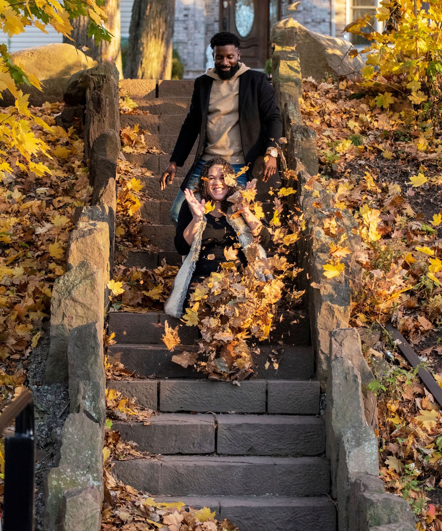A couple during their maternity shoot has some fun and the father throws a pile of leaves onto the mom to be as she laughs in Nutley NJ