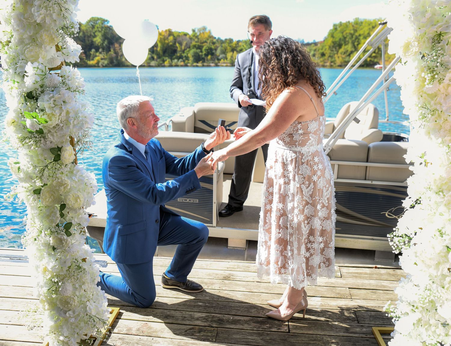 A man is on one knee giving a ring to his wife at their vow renewal ceremony on a boat dock on a lake in Sparta, NJ