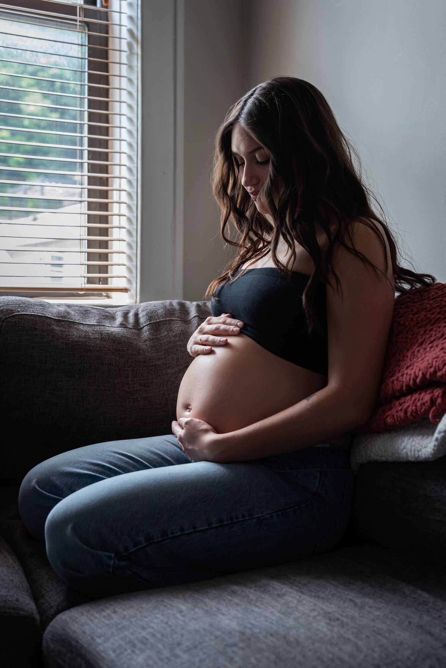 A pregnant woman sits on her couch holding her belly looking down at her stomach during her maternity session in Edgewater, NJ
