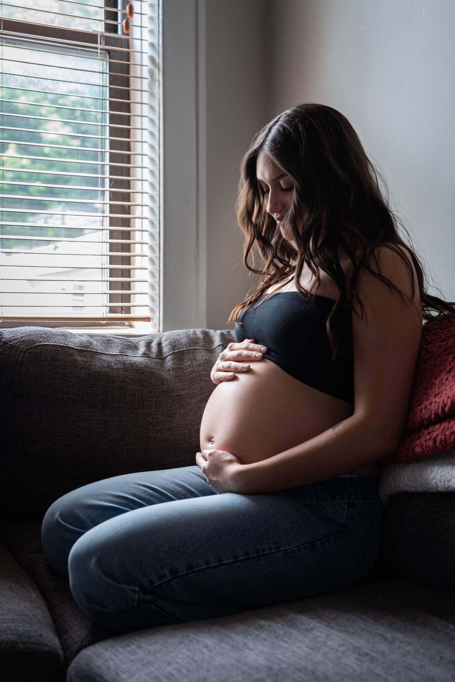 A pregnant woman sits on her couch holding her belly looking down at her stomach during her maternity session in Edgewater, NJ