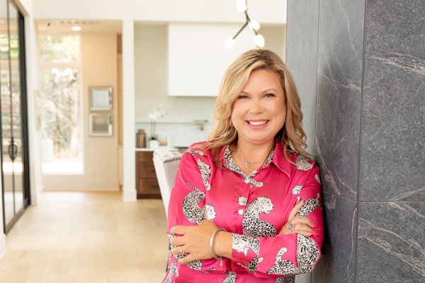 A cheerful blonde woman in a pink floral blouse leans against a black marble wall in a modern home.