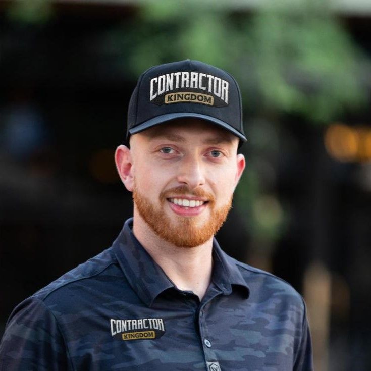 Young man with red beard wearing Contractor Kingdom branded hat and polo shirt, smiling outdoors.