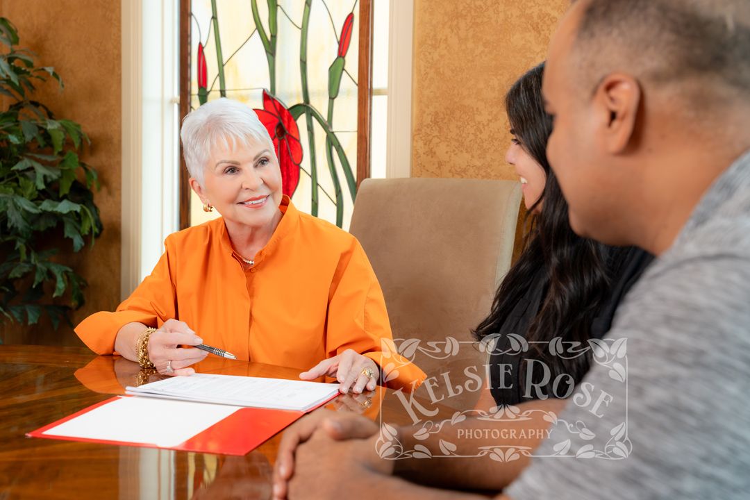 Elderly woman in orange blouse signing documents at a table with a couple during a professional meeting.