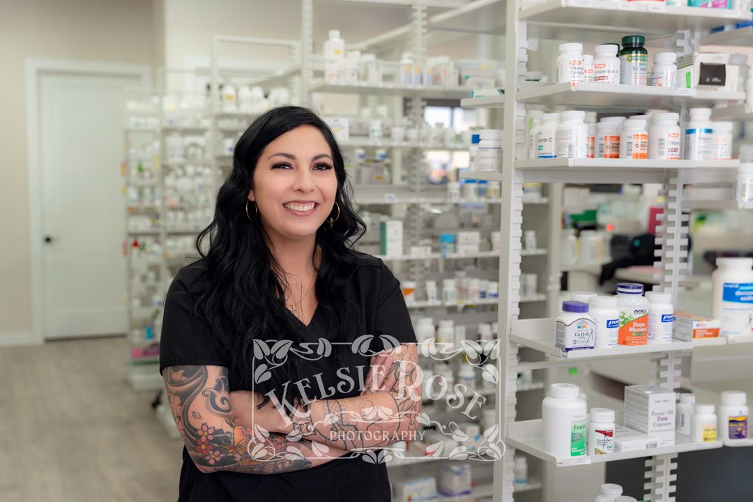 Female pharmacy technician in black scrubs smiling in front of medication shelves.