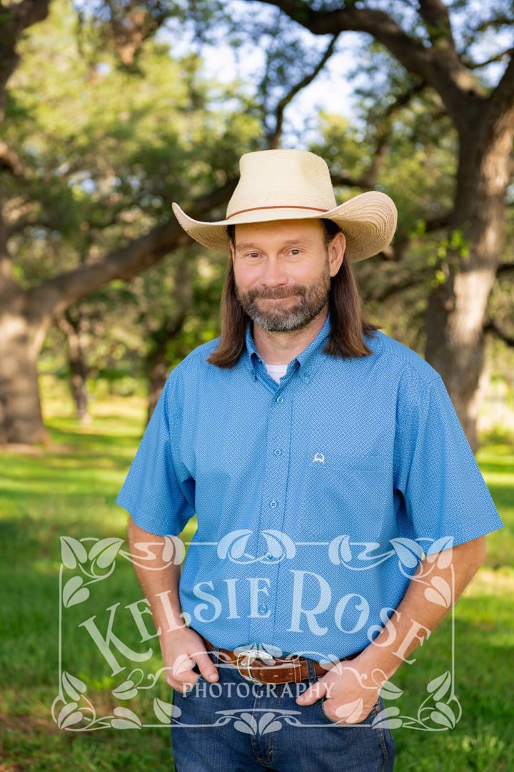 Professional portrait of smiling man with long hair in straw cowboy hat and blue shirt posing outdoors under oak trees.